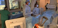 An interventional radiology physician and resident in surgical gowns perform live fluoroscopic imaging. A technologist prepares the sterile procedure table in the background. 