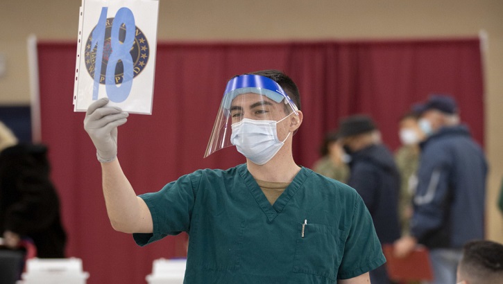 Image of Military health personnel wearing a mask and a face shield holding up a sign that has the number eighteen on it .