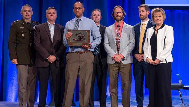 Image of Dr. Sumil Shah accepts the Outstanding Program Management (Team) Award, along with some members of his team from Ms. Seileen Mullen, the acting secretary of Defense for Health Affairs. MHSRS provides a collaborative setting for the exchange of information between military providers with deployment experience, research and academic scientists, international partners, and industry on research and related health care initiatives, such as Combat Casualty Care, Operational Medicine, Clinical and Rehabilitative Medicine, Medical Simulation and Information Sciences, and infectious Diseases. .